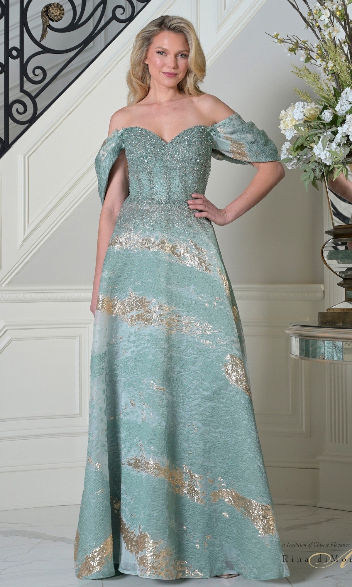 A woman is indoors wearing the Rina Di Montella RD3405 long formal dress in seafoam, featuring a fitted beaded bodice, metallic gold accents, and an elegant A-line organza skirt.